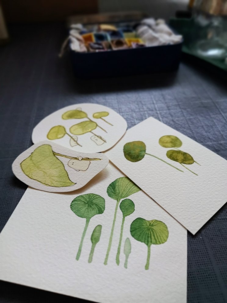Des mini-illustrations à l'aquarelle, sur le thème de la couleur verte, posées
sur un plan de travail. Les illustrations représentent des feuilles de plantes
imaginaires et des petits êtres de la nature. En arrière-plan, on peut voir une
petite boîte avec des godets d'aquarelle.