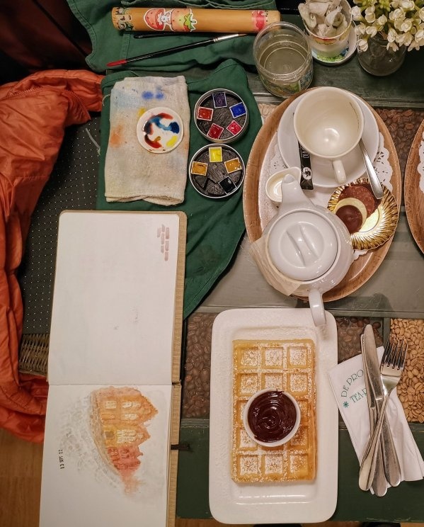 Vue de dessin d'une table dans un café. On peut voir une théière, des
    couverts, un tasse et une assiette avec une gaufre et son coulis de
    chocolat. À côté, des godets d'aquarelle et un carnet avec une esquisse
    des façades de briques de Bruges.