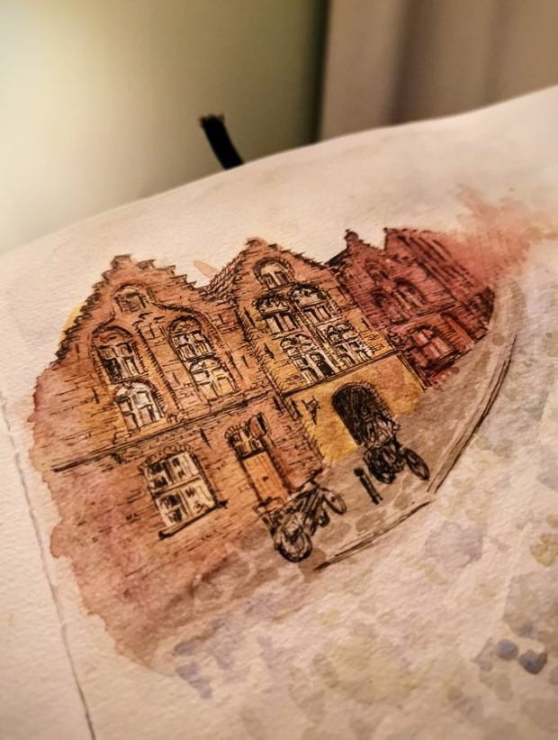 Gros plan sur un carnet de dessin. On peut voir une esquisse à
l'aquarelle d'une façade de briques dans le style de Bruges avec une rue
pavée et la rue qui se fond dans la brume en arrière-plan.