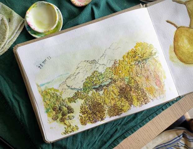 Un carnet de dessins ouvert posé sur un plan de travail avec des coupelles
tachées d'aquare. Une illustration est peinte sur la page ouverte du carnet
et représente un paysage de montagne avec des arbres à l'automne. Il est daté
au 25 septembre 2022.