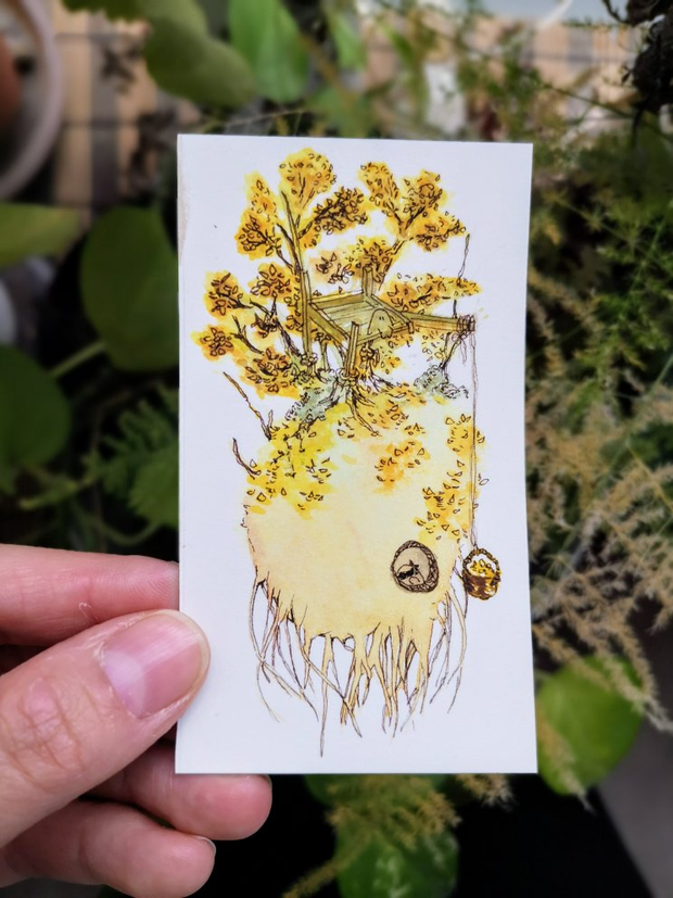 Une main qui tient une mini-illustration à l'aquarelle. L'aquarelle représente
une boule de terre aux couleurs automnales avec un arbre aux feuilles dorées
dessus. Des petits êtres de la forêt habitent sur cette boule de terre.