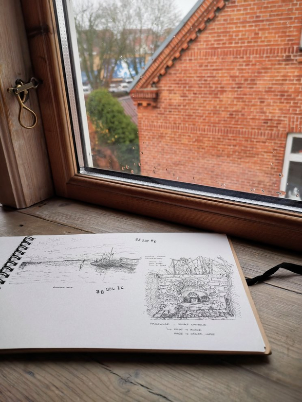 Un carnet de dessin ouvert sur le rebord d'une fenêtre. Le carnet présente
deux dessins à l'encre. L'un représente une étendue d'eau avec un bateau
amarré. L'autre une fontaine dans un mur en pierre. À travers la fenêtre, on
peut une partie d'une façade de maison en briques rouges.
