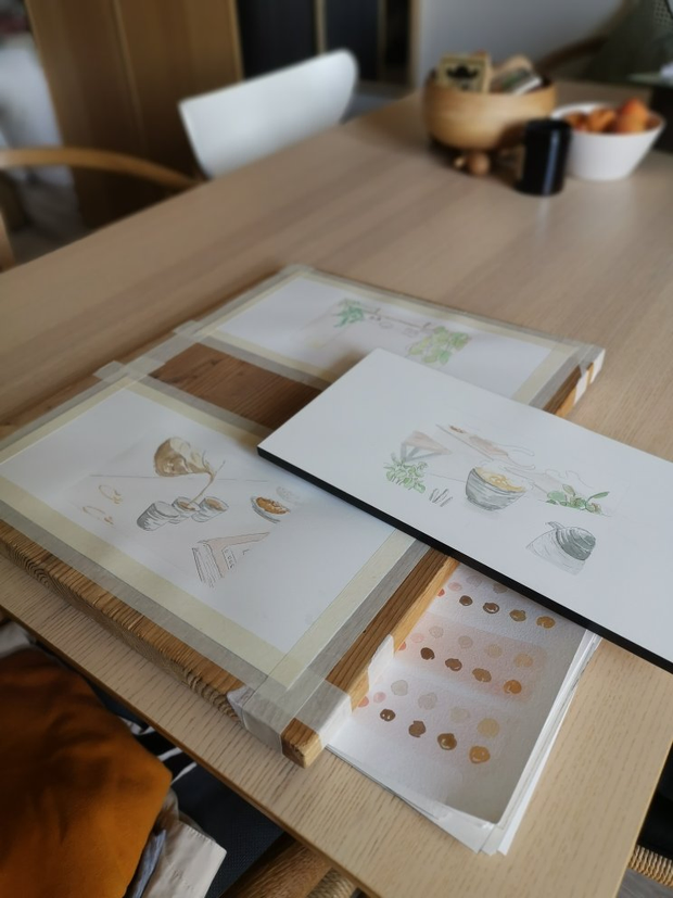 Une table sur laquelle sont posées plusieurs illustrations en cours à
l'aquarelle. Les illustrations représentent des tasses de café et des plantes.
On peut également voir un nuancier posé à côté.
