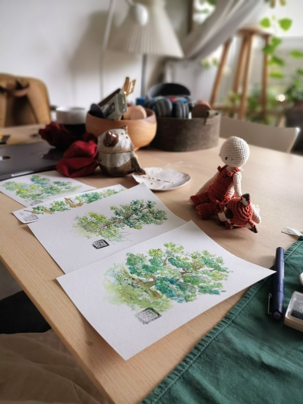 Une table sur laquelle sont posées des doudous faits en crochet et des
illustrations à l'aquarelle. Les illustrations représetent des arbres
verdoyants. En arrière-plan, pleins d'objets en vrac encombrent le bout de la
table.
