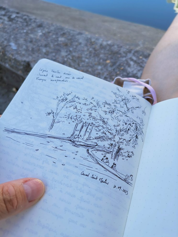 Une main qui tient un croquis fait au stylo à encre. L'illustration représente
des arbres autour d'un canal avec des gens assis sur le rebord.
