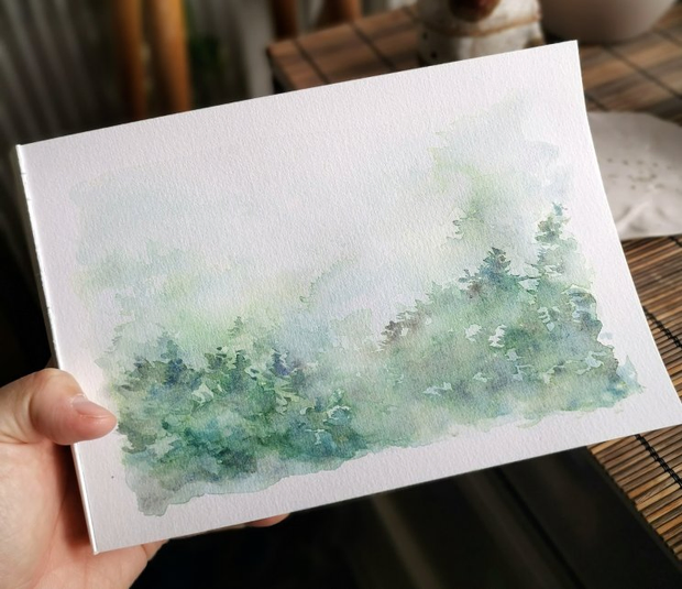 Une main qui tient un illustration à l'aquarelle. L'illustration représentent
une forêt en hiver dans une montagne embrumée.
