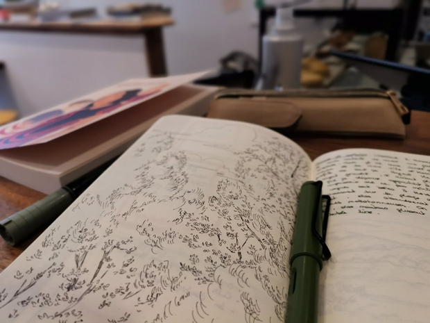 Gros plan sur une page d'un carnet sur laquelle on voit une illustration
d'arbres au stylo noir. En arrière-plan, sont posés sur la même table, un
livre et des stylos.
