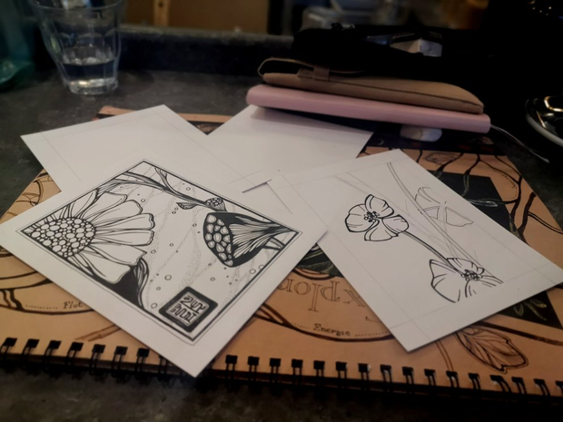 Deux illustrations à l'encre qui représentent des fleurs et des plantes
imaginaires. On peut voir des feuilles et un carnet rose posés en
arrière-plan.

