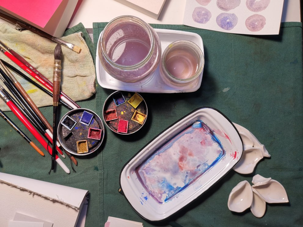 Table de travail vue du dessus avec posées en vrac, des coupelles en
porcelaine, des godets d'aquarelle, des pinceaux et deux bocaux d'eau.
