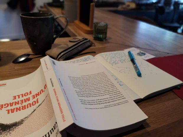 Gros plan sur un livre posé à l'envers sur le comptoir d'un café. On peut voir
en-dessous un carnet de notes ouvert et un stylo bleu. À côté, un grand mug
noir.
