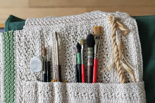 Vue du dessus d'une pochette de pinceaux. La pochette est faite en crochet,
d'une couleur blanc cassé. On distingue plusieurs types de pinceaux ainsi
qu'un petite corde tressée dedans.
