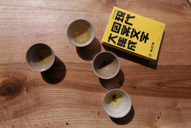 Vue du dessus d'une table sur laquelle sont posées plusieurs petites tasses en
céramique grise ainsi qu'un petit livre à la couverture jaune avec de grosses
inscriptions en caractères japonais.
