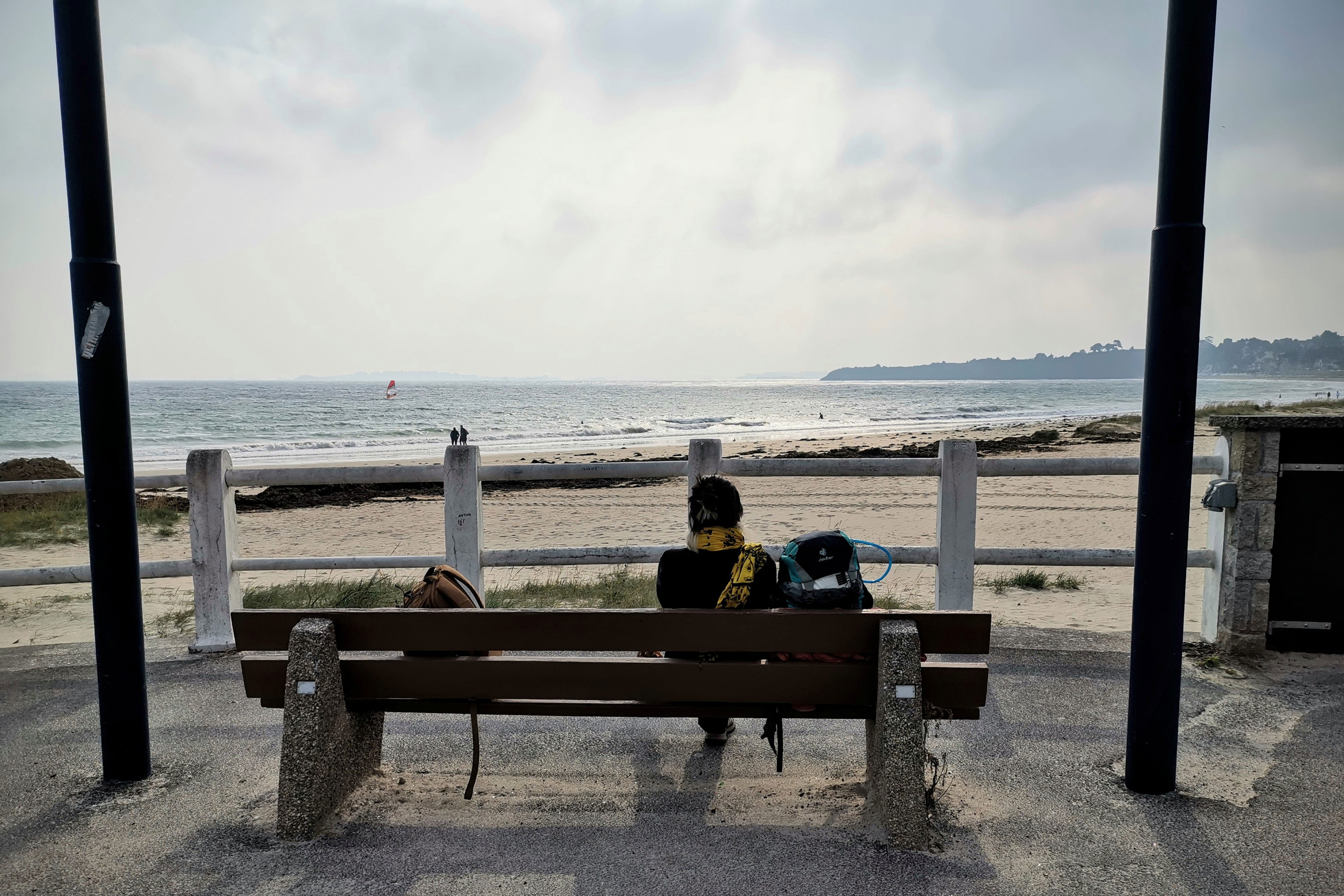 Une femme de dos assise sur un banc en face d'une mer calme. Un grand
    ciel blanc est au-dessus d'elle et de ses deux sacs à dos.